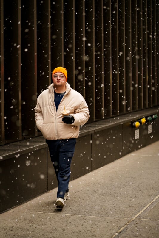 A man wearing a beige puffer jacket, orange beanie, and navy pants walks down a city sidewalk in the snow, holding a drink and looking ahead. Snowflakes fall around him and a dark building lines the street.