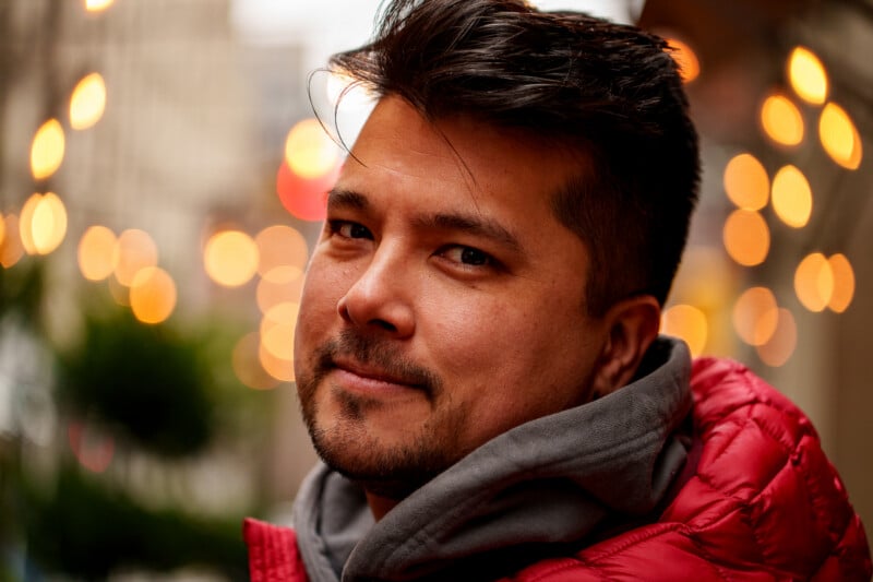A man with short dark hair and a trimmed beard, wearing a red jacket and gray hoodie, looks at the camera with a slight smile. Warm, blurred lights decorate the background, giving a cozy city atmosphere.
