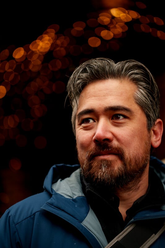 A man with gray-streaked hair and a beard looks to the side, wearing a blue jacket. Warm, out-of-focus lights create a bokeh effect in the dark background.