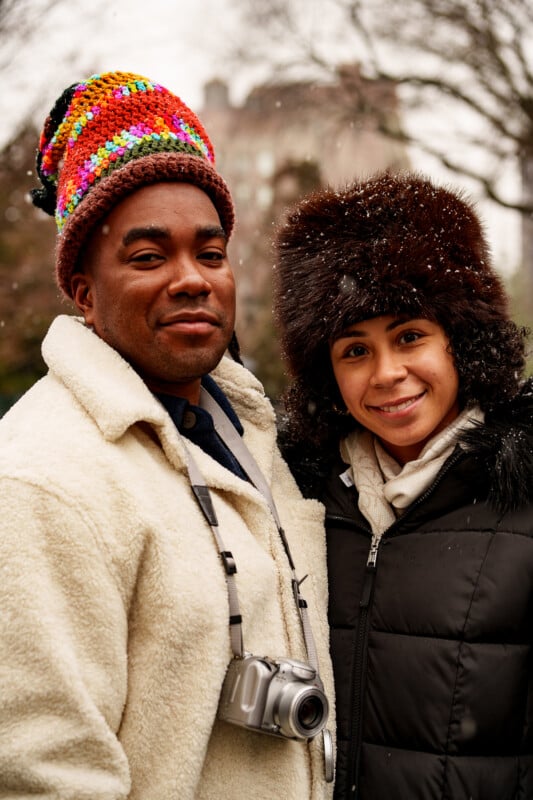 A man and woman stand outdoors in winter clothing. The man wears a colorful knit hat, cream jacket, and has a camera around his neck. The woman wears a furry hat and black coat. Snow is falling and trees are visible in the background.