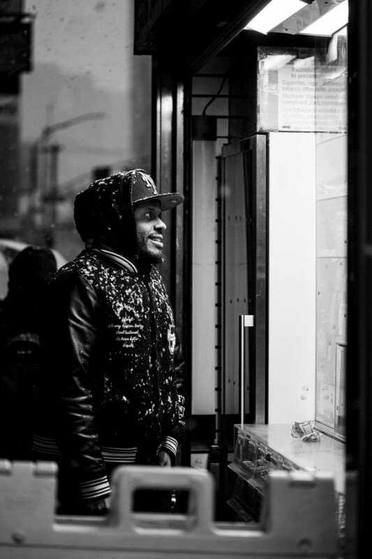 A man wearing a hooded jacket and cap, dusted with snow, smiles while standing at a counter inside a store. The scene is captured in black and white through a window.