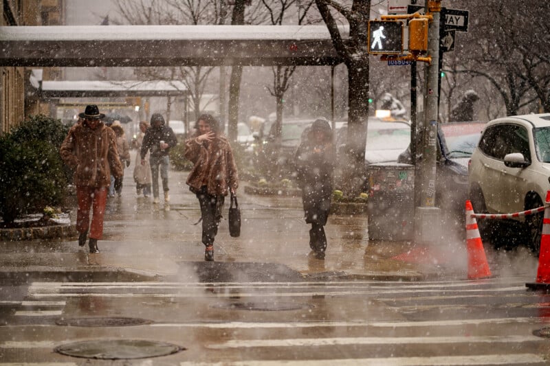 People cross a city street in heavy snow, shielding themselves from the falling snowflakes. Traffic cones and wet pavement are visible, and trees line the sidewalk as cars are parked along the road.
