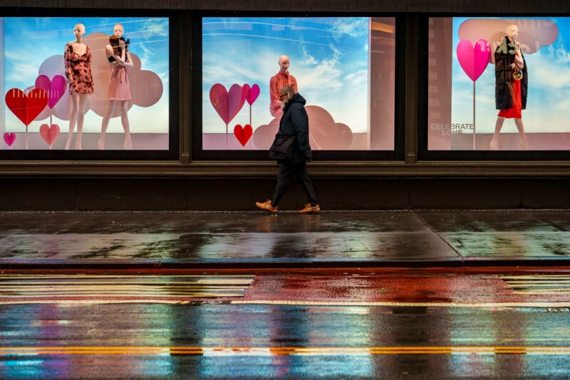 A person walks past store windows with mannequins dressed in red and pink outfits, surrounded by heart decorations. The wet street reflects the display and the person, suggesting a recent rain.