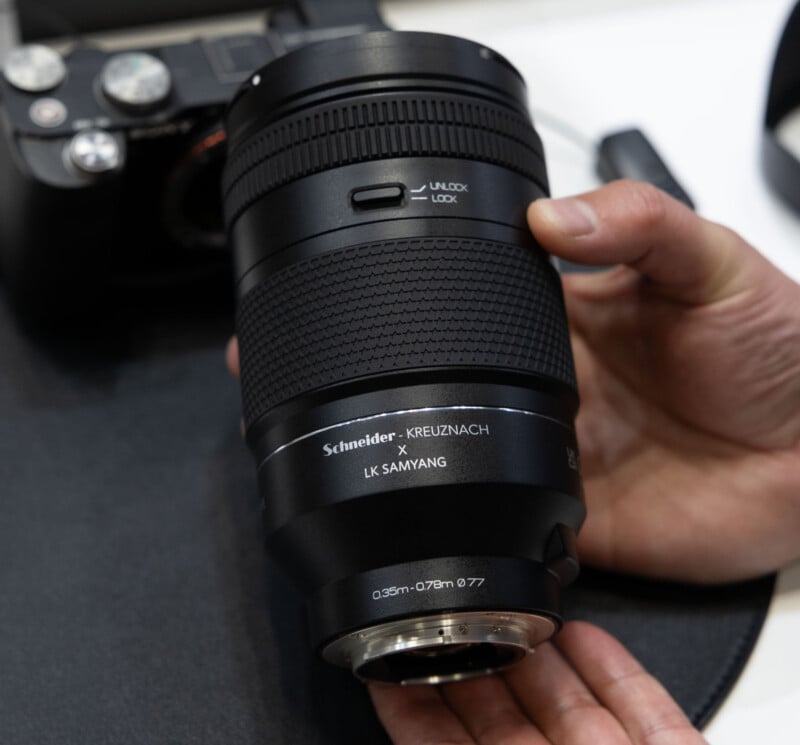 A person holds a Schneider-Kreuznach x LK Samyang camera lens, with a camera body and strap visible in the background. The lens features a textured grip and a lock/unlock switch.
