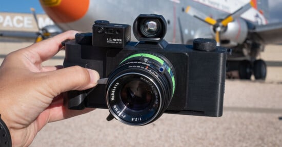 A hand holds a black film camera with a large lens and a viewfinder, outdoors in front of a blurred airplane and the word "SPACE" partially visible on a building in the background.