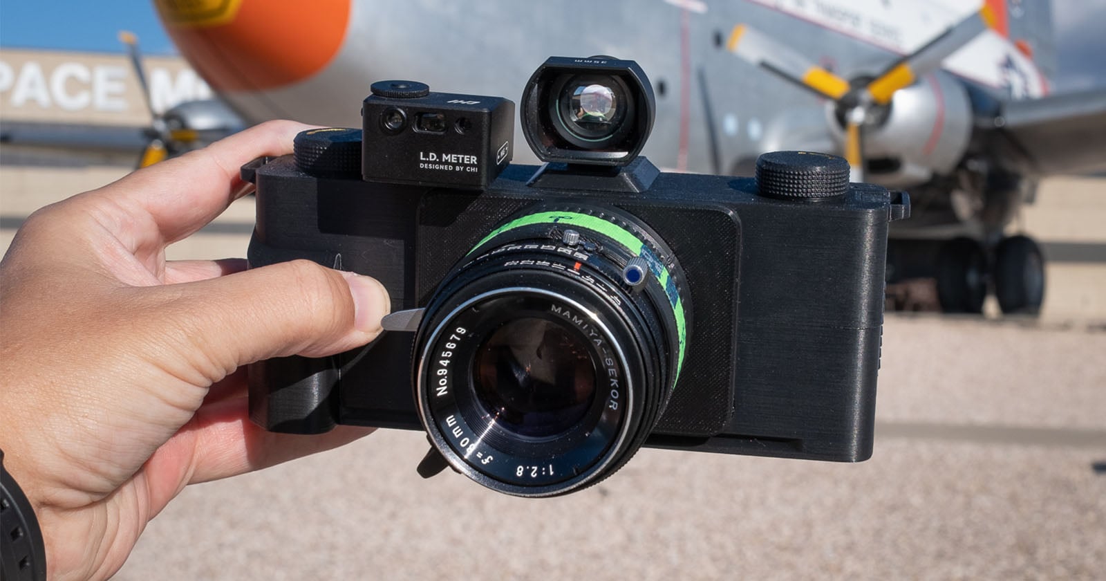 A hand holds a black film camera with a large lens and a viewfinder, outdoors in front of a blurred airplane and the word "SPACE" partially visible on a building in the background.