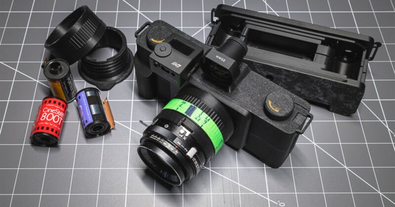 A black film camera with a green label is placed on a cutting mat alongside rolls of 35mm film, lens adapters, and an open camera back.