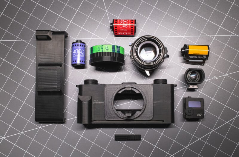 Various camera parts, film rolls, and lenses are arranged neatly on a grid-patterned surface, including a black film camera body, viewfinder, lens, 35mm film canisters, and other small camera components.