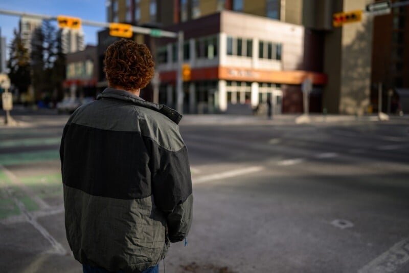 Un hombre con pelo rojo rizado y una chaqueta gris-negra se encuentra en un paso de peatones de la ciudad, de espaldas a la cámara, con edificios y semáforos visibles al fondo.