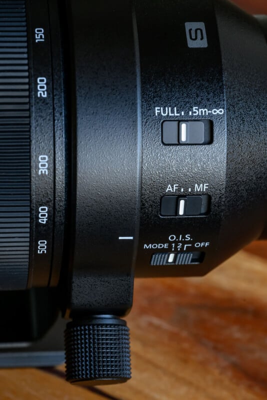 Close-up of a camera lens showing three switches labeled "FULL .5m–∞," "AF–MF," and "O.I.S. MODE 1/2/OFF," with part of the lens barrel and a textured adjustment knob visible.