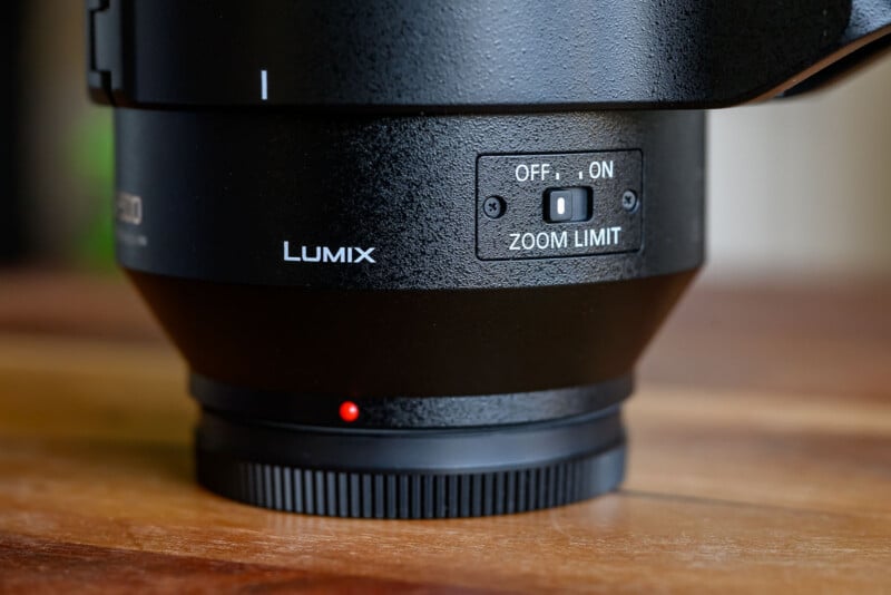 Close-up of a black Lumix camera lens showing a switch labeled "ZOOM LIMIT" with options for "OFF" and "ON," sitting on a wooden surface. A small red dot is visible near the lens mount.