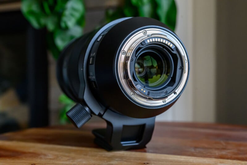 Close-up of the rear mount of a large camera lens resting on a wooden surface, with green leaves blurred in the background.