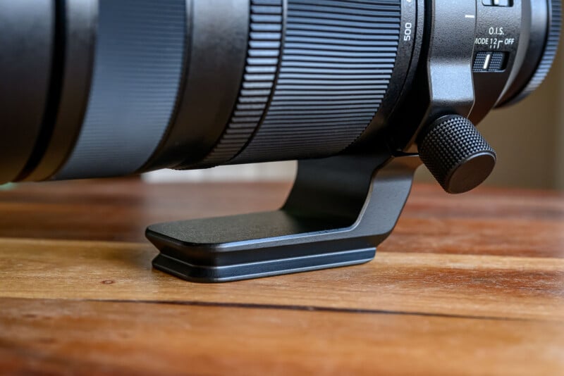 Close-up of a camera telephoto lens with a tripod mounting collar, resting on a wooden surface.