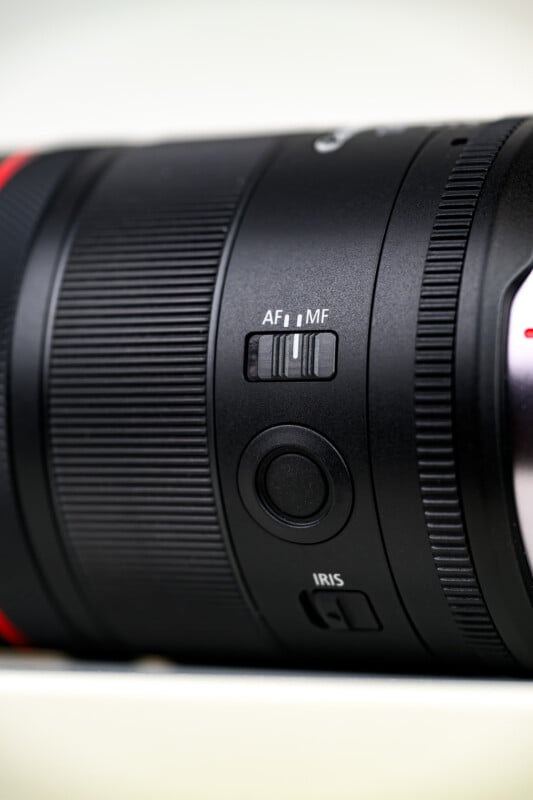 Close-up of a camera lens showing a textured grip, an AF/MF (autofocus/manual focus) switch, a round customizable button, and an iris control switch on the lens barrel.