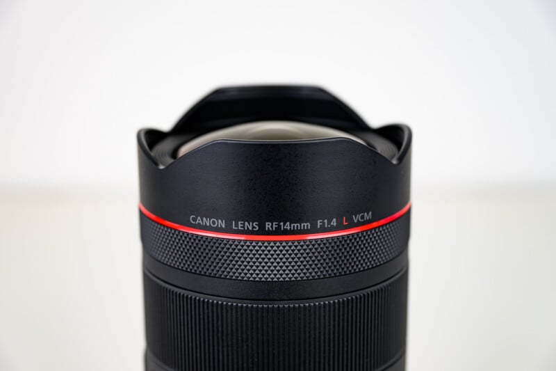 Close-up of a Canon camera lens labeled "Canon Lens RF14mm F1.4 L VCM" against a plain, light background. The lens has a textured grip and a distinctive red ring near the top.