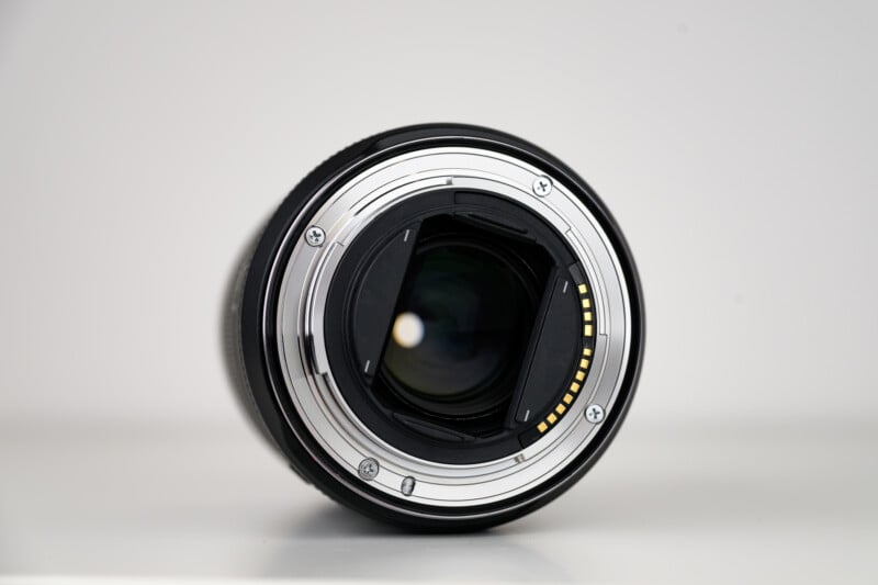 Close-up view of the rear side of a camera lens, showing the metal mount, electronic contacts, and internal lens elements against a white background.