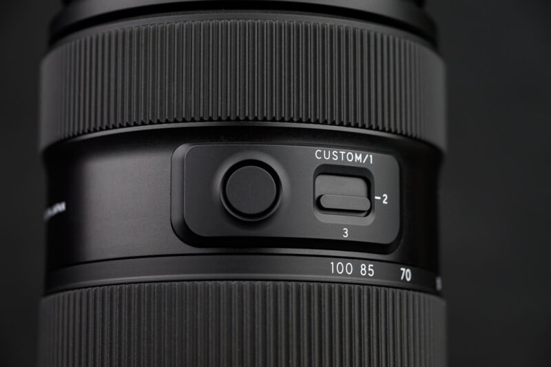 Close-up of a camera lens showing a control panel with a circular button, a sliding switch labeled "CUSTOM/1" with settings 1, 2, and 3, and focal length markings from 70 to 100. The lens has a textured grip.