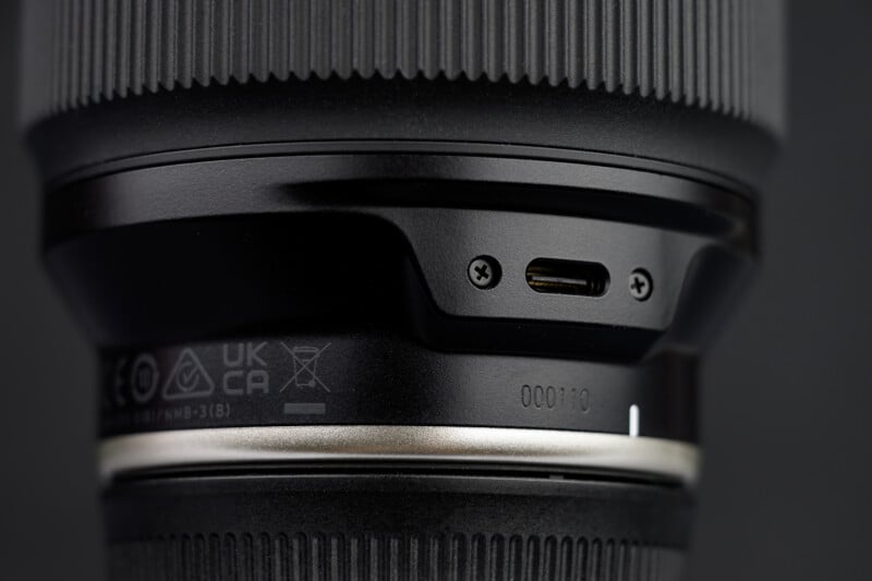 Close-up of a camera lens showing a USB-C port, small screws, and certification markings, with textured focus and zoom rings visible in the background.