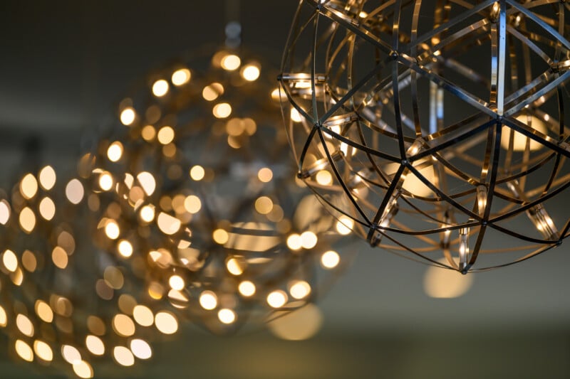 Close-up of modern spherical metal light fixtures with warm, glowing bulbs inside. The background shows blurred lights, creating a soft bokeh effect with circular patterns.