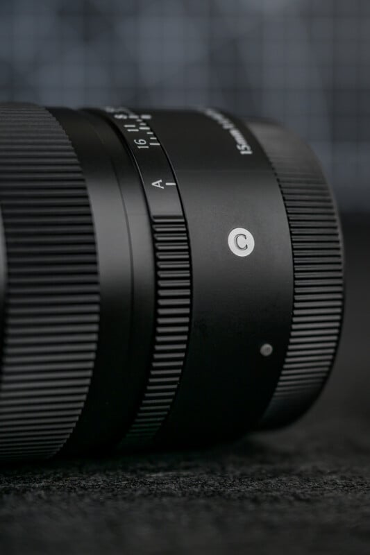 Close-up of a black camera lens lying on its side, showing a focus ring with textured grip, aperture markings, and a “C” symbol on a matte background. The background is blurred with a grid pattern.