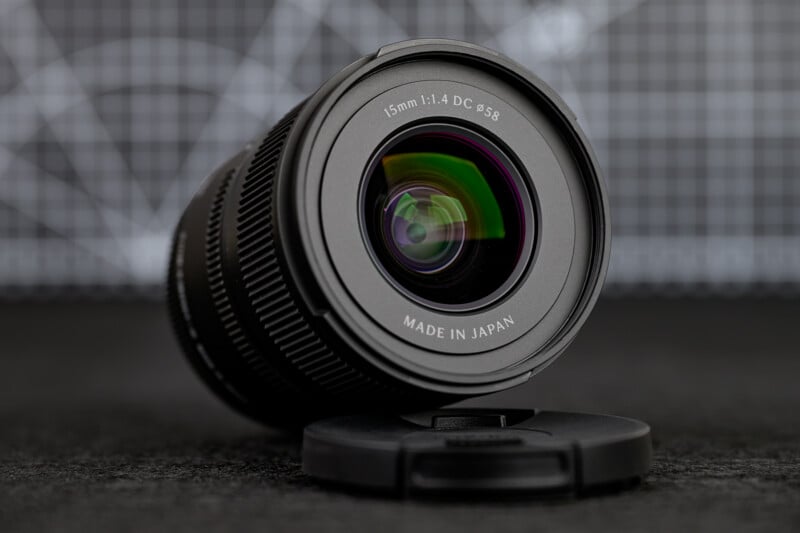 1.4 DC ϕ58 MADE IN JAPAN," with its lens cap lying in front on a dark surface and a grid-patterned background.