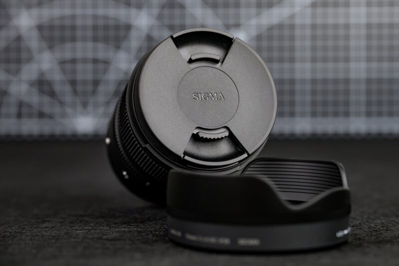 A Sigma camera lens with the lens cap on is positioned upright, next to a detached lens hood, on a dark textured surface with a graph grid background.