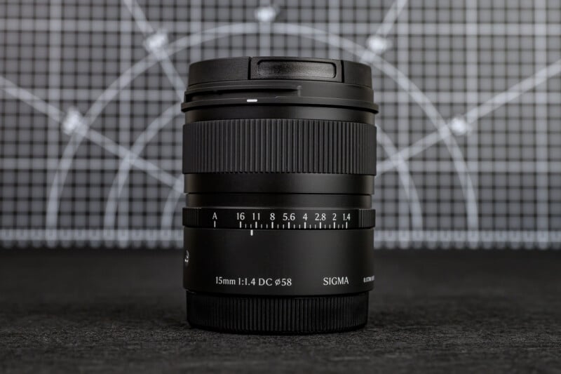 A Sigma 15mm f/1.4 DC camera lens stands upright on a dark surface, with a grid and circular pattern in the blurred background. The aperture and focus markings are visible on the lens barrel.