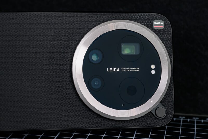 Close-up of a smartphone’s rear camera module with three lenses, LED flash, and Leica branding, set within a textured black surface and a silver ring, on a checkered background.