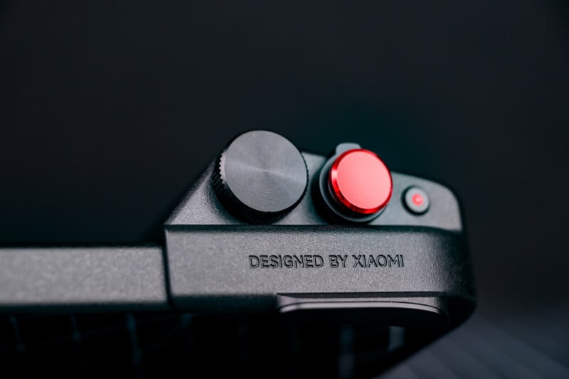 Close-up of a black electronic device with a large black knob, a red button, and a small power button. The text "DESIGNED BY XIAOMI" is engraved on the device’s surface.