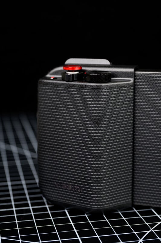 A close-up of a black textured camera grip with a red button and a black dial, set against a black background with a white grid pattern.