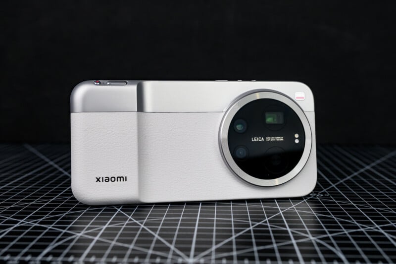 A silver Xiaomi smartphone with a large round Leica-branded camera module on the back, resting on a black grid-patterned surface with a dark background.