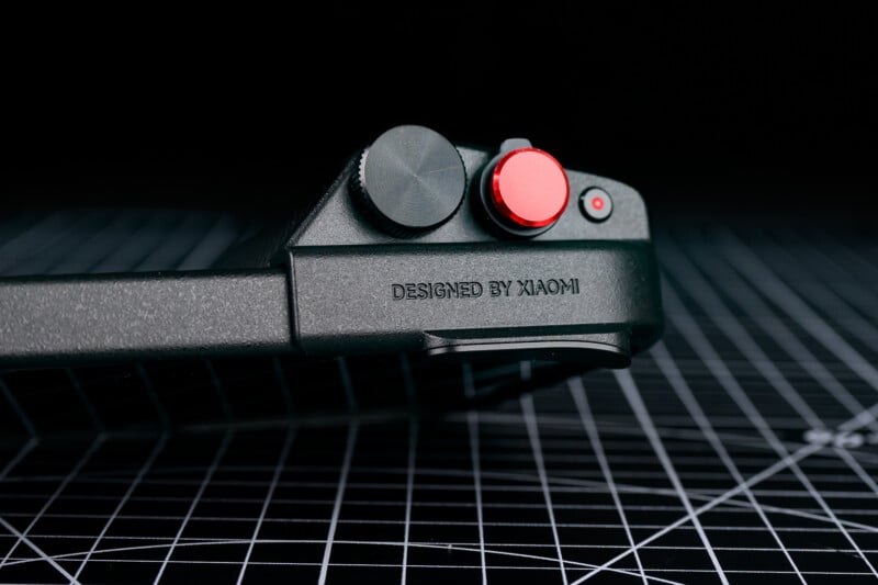 Close-up of a black gaming controller with a red joystick and button, featuring the text "DESIGNED BY XIAOMI" on its side, resting on a grid-patterned black surface.