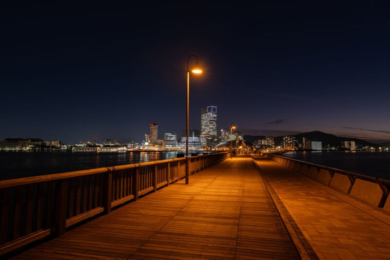 A well-lit wooden pier extends into the water at night, leading toward a distant city skyline with illuminated modern buildings and a dark, clear sky above.