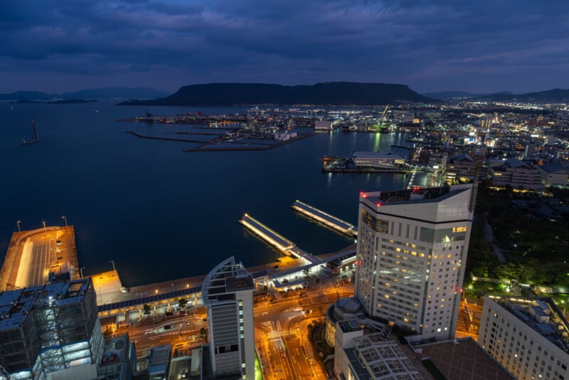 A cityscape at dusk with illuminated buildings and roads by the waterfront. The harbor extends into calm water, with distant mountains and a darkening sky in the background.