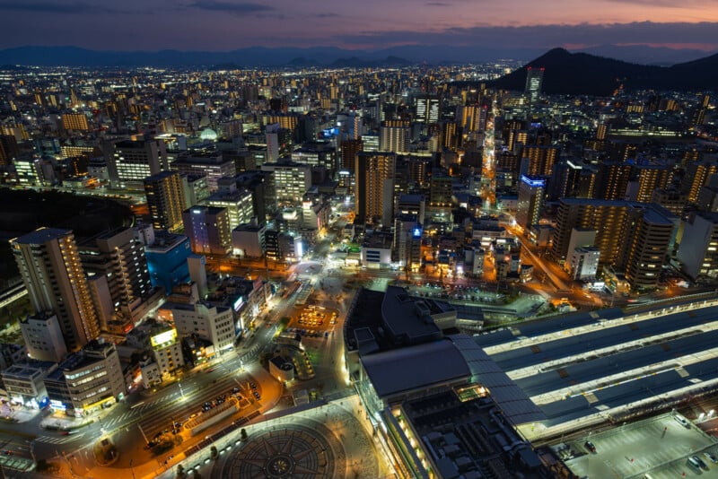 A cityscape at dusk with numerous illuminated buildings, roads, and a visible train station in the foreground. The sky is darkening, and mountains are faintly visible on the horizon.