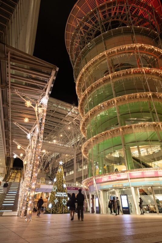 Plaza de la ciudad moderna por la noche decorada con luces navideñas. Un árbol de Navidad gigante y resplandeciente se encuentra cerca de un edificio alto de vidrio, y la gente camina bajo esculturas de renos resplandecientes y cadenas de luces navideñas.