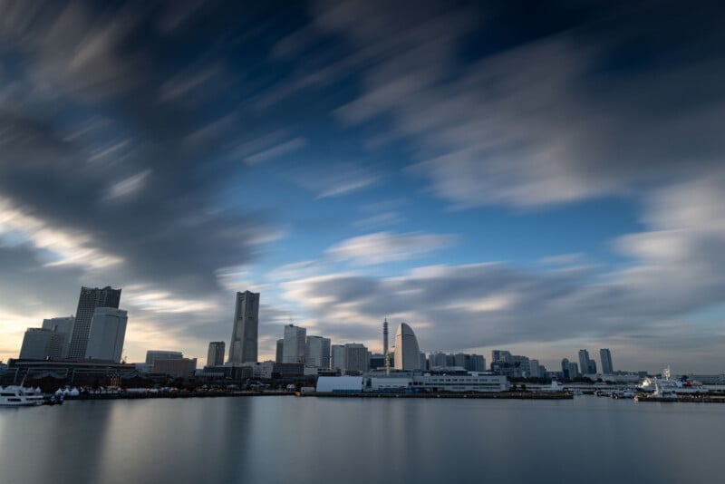 A city skyline with tall modern buildings stands along a calm waterfront under a dramatic sky, with long, blurred clouds moving across at sunset or sunrise. The water reflects part of the skyline and boats are docked near the shore.