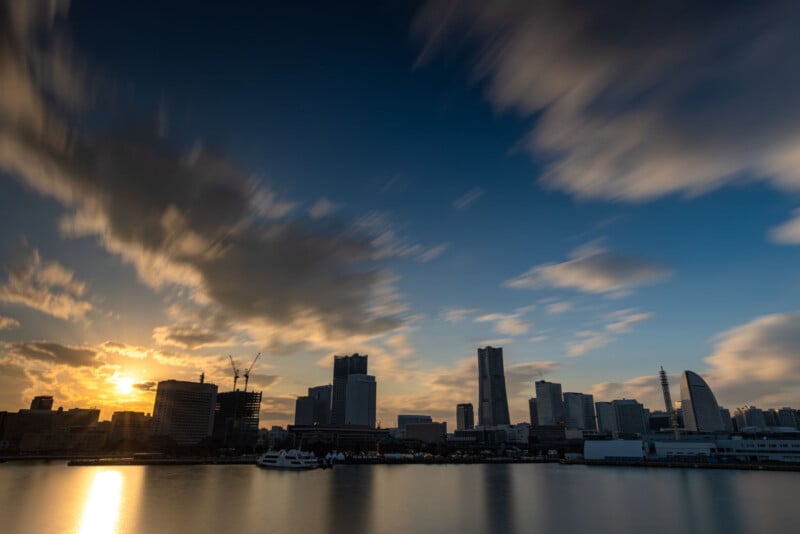 City skyline at sunset with tall buildings silhouetted against a dramatic sky, clouds streaked by long exposure, and sunlight reflecting off calm water in the foreground.
