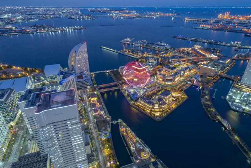 Aerial view of Yokohama at dusk, featuring illuminated city buildings, a colorful Ferris wheel, and a harbor with ships, all reflecting on the water under a blue evening sky.