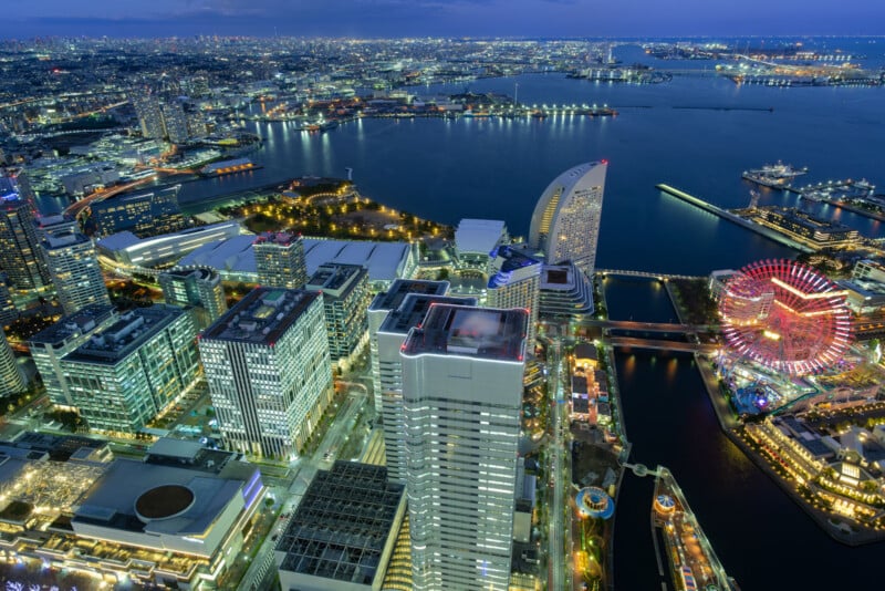 Vista aérea de Yokohama, Japón al atardecer, que muestra rascacielos iluminados, bahía con puente, gran noria y luces de la ciudad reflejadas en el agua.