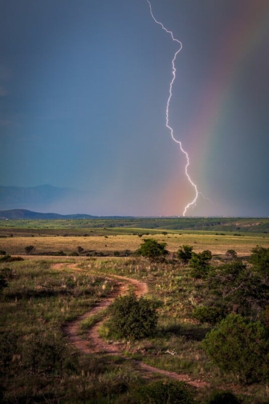 A vivid lightning bolt strikes the ground under a dark sky with a faint rainbow, above a winding dirt path running through a grassy, open landscape with distant hills.