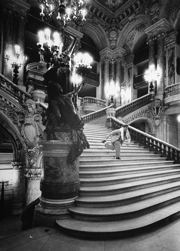 Black and white photo of a grand, ornate staircase with chandeliers and elaborate carvings; a person in mid-leap dances up the steps, creating a dynamic contrast with the elegant, historic architecture.