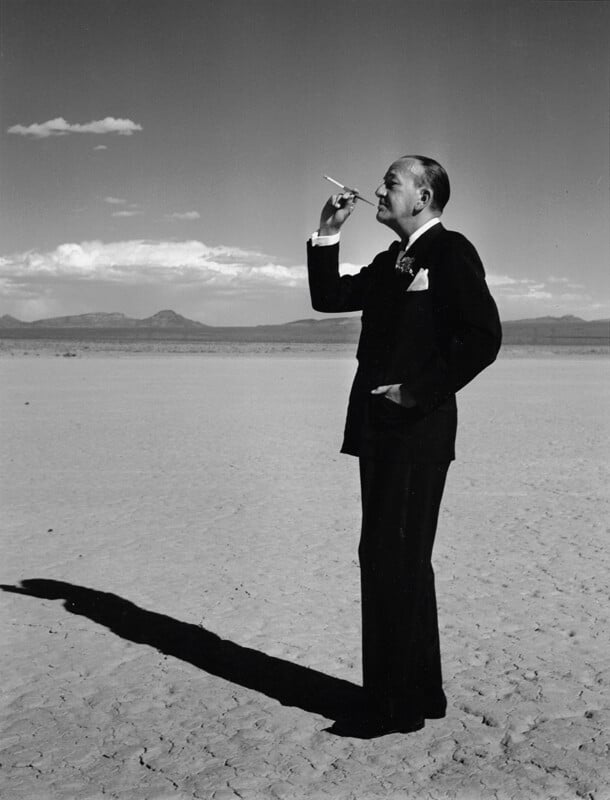 A man in a suit stands on cracked desert ground, holding a cigarette holder to his mouth. He looks thoughtfully ahead, with one hand in his pocket. His long shadow stretches across the sunlit landscape under a mostly clear sky.