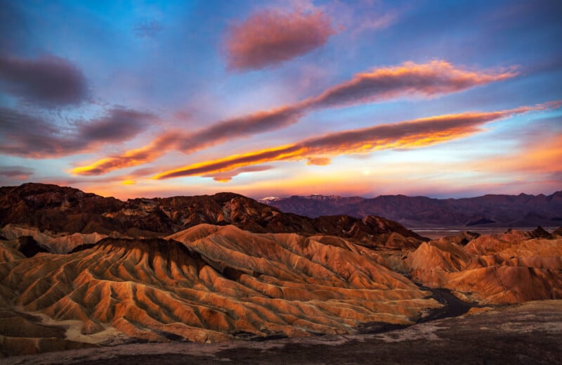 Colorful sunset sky with streaked clouds over rugged, golden-brown hills and mountains in a desert landscape. The warm light highlights the ridges and textures, creating a dramatic, serene scene.