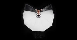 A person stands behind a large, open, white photography umbrella with only their head and hands visible above it, against a black background.