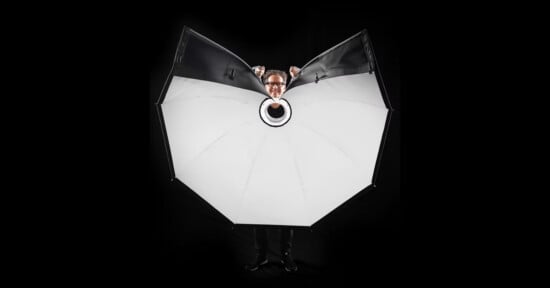 A person stands behind a large, open, white photography umbrella with only their head and hands visible above it, against a black background.