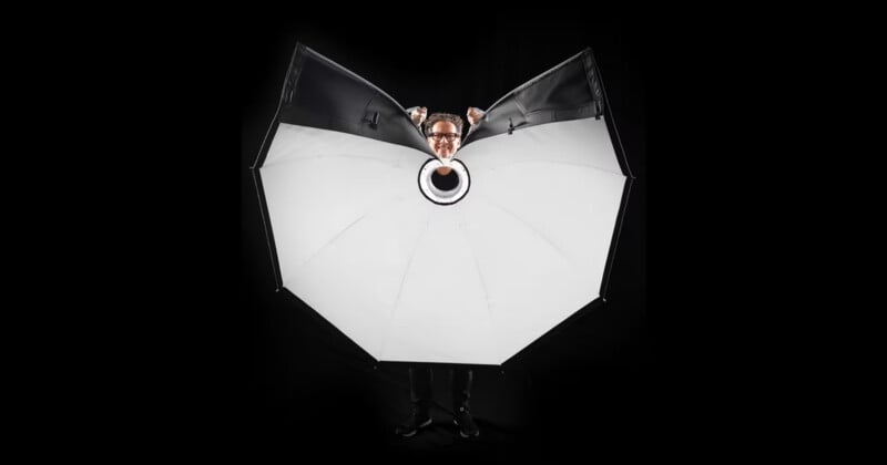 A person stands behind a large, open, white photography umbrella with only their head and hands visible above it, against a black background.