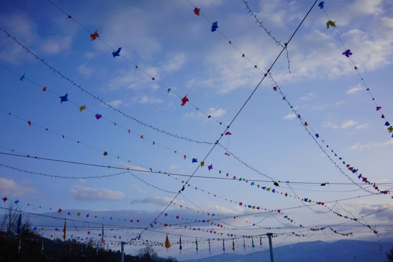 El cielo exterior está lleno de coloridos pájaros de papel y luces de cadena, creando una atmósfera festiva contra el cielo azul y las nubes blancas. Los árboles y las montañas distantes son claramente visibles.