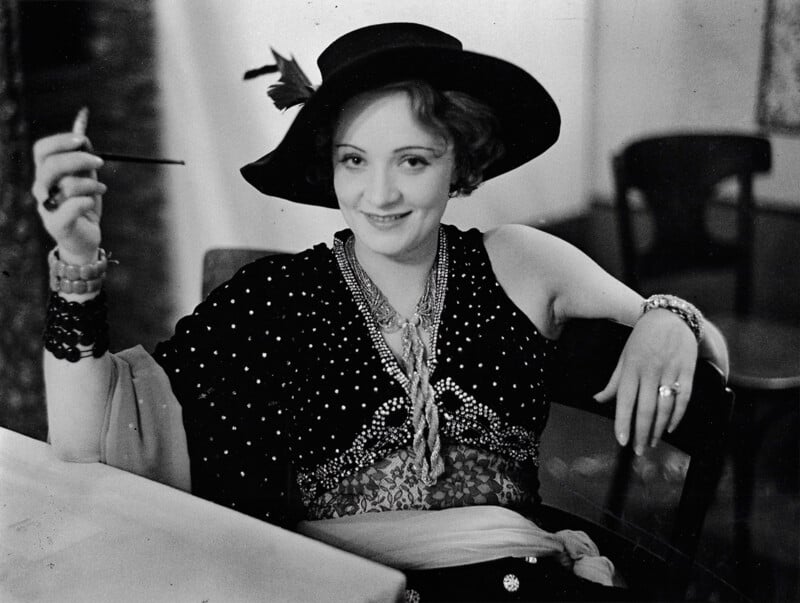 A woman wearing a wide-brimmed hat and vintage polka-dot dress smiles at the camera while holding a cigarette in a long holder, seated at a table with jewelry and accessories.