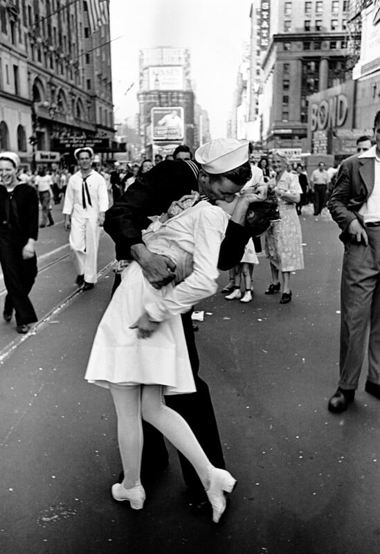 A sailor passionately kisses a woman in a white dress amid a crowd in Times Square, New York City, celebrating the end of World War II. People around them smile, walk, and observe the joyful scene.